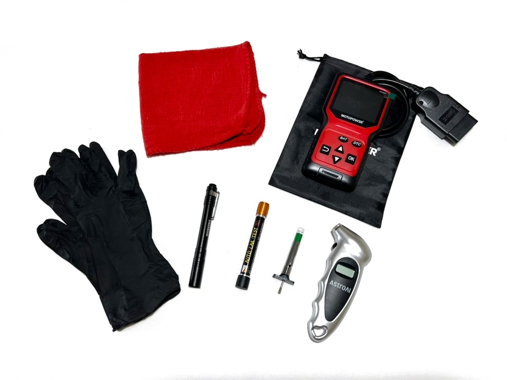 The hands-on tools from the kit laid out on a white surface: red shop rag, black nitrile gloves, pen flashlight, paint thickness gauge, tread depth gauge, digital tire pressure gauge and an OBD2 scanner on its pouch